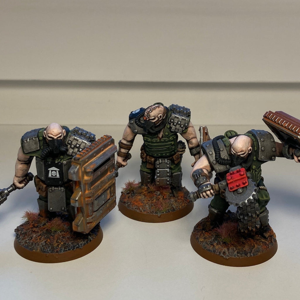 Three painted fantasy warrior miniatures on round bases, posed against a plain light gray background. The figures are muscular and heavily armored in dark green, gray, and brown tones, with one holding a chainsword and the others carrying shields or weapons.